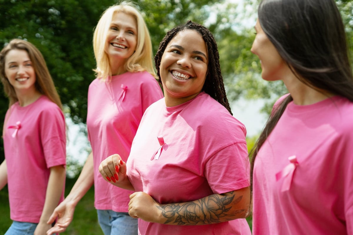 Four smiling women of diverse ages and ethnicities walk outdoors, wearing pink shirts adorned with pink ribbons, symbolizing their support for breast cancer awareness and research