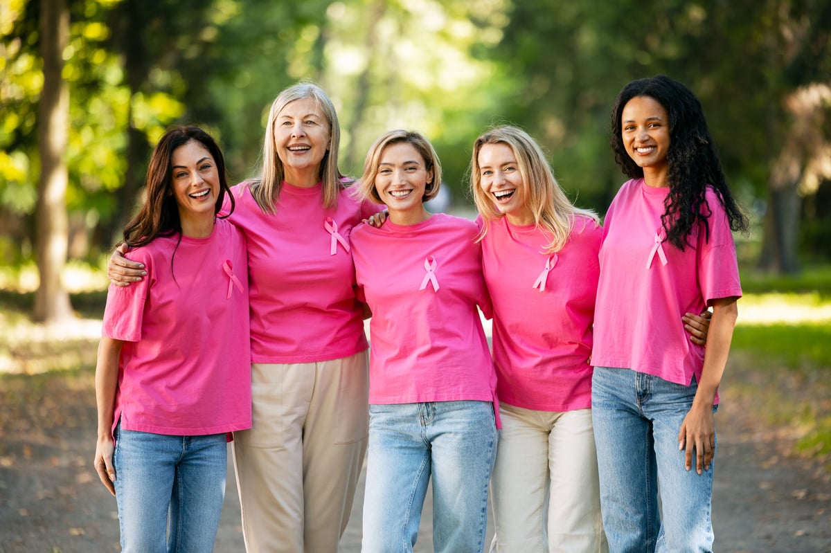 Group of smiling women wearing pink ribbons and t-shirts, standing together in a park, showing solidarity for breast cancer awareness